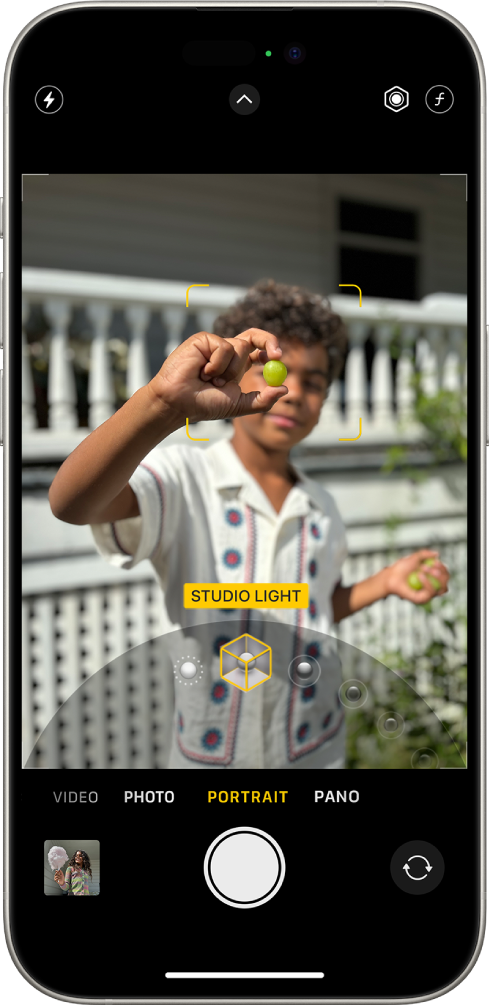 Kamera-skjermen i Portrett-modus. I søkeren er motivet skarpt, og bakgrunnen er uskarp. Hjulet for å velge Portrettlyssettinger er åpent nederst i bildet, og Studiolys er valgt. Øverst til venstre på skjermen er Blits-knappen, øverst i midten er Kamerakontroller-knappen, og øverst til høyre er knappene for å justere intensitet for Portrettlyssetting og Dybdekontroll. Nederst på skjermen er, fra venstre til høyre, Bilde- og Videovisning-knappen, Ta bilde-knappen og Kameravelger-knappen.