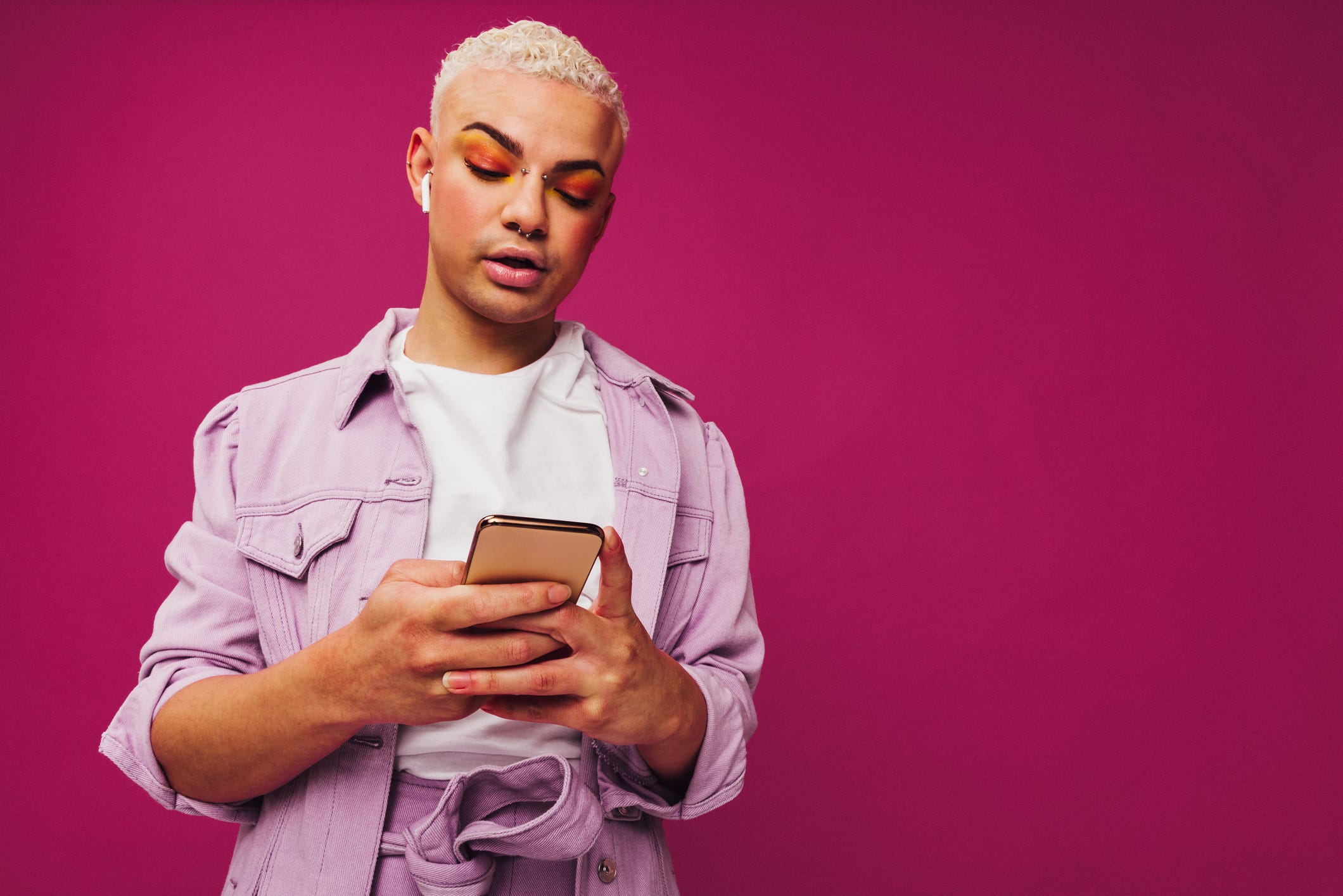 browsing trendy podcasts stylish young man using a smartphone while wearing wireless earphones in a studio non conforming queer man standing alone against a purple background