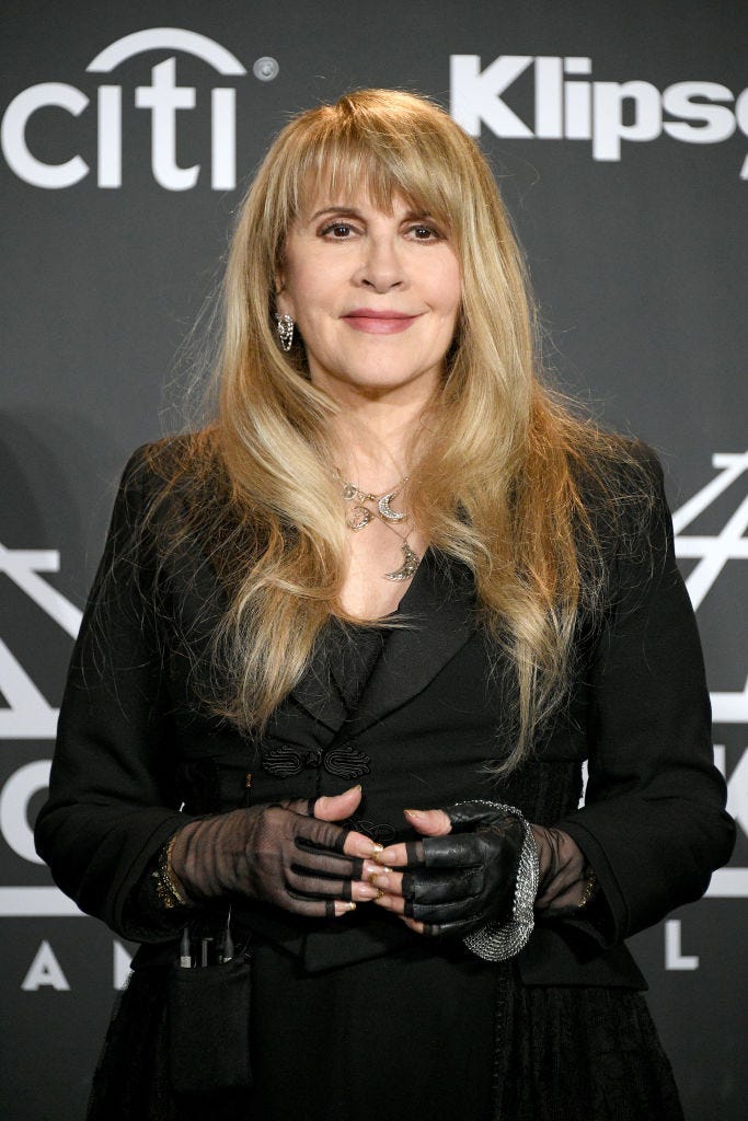 stevie nicks smiles and looks just past the camera while standing in front of a dark gray backdrop, she wears all black, several silver and gold necklaces, and large dangling earrings, her long blond hair is down