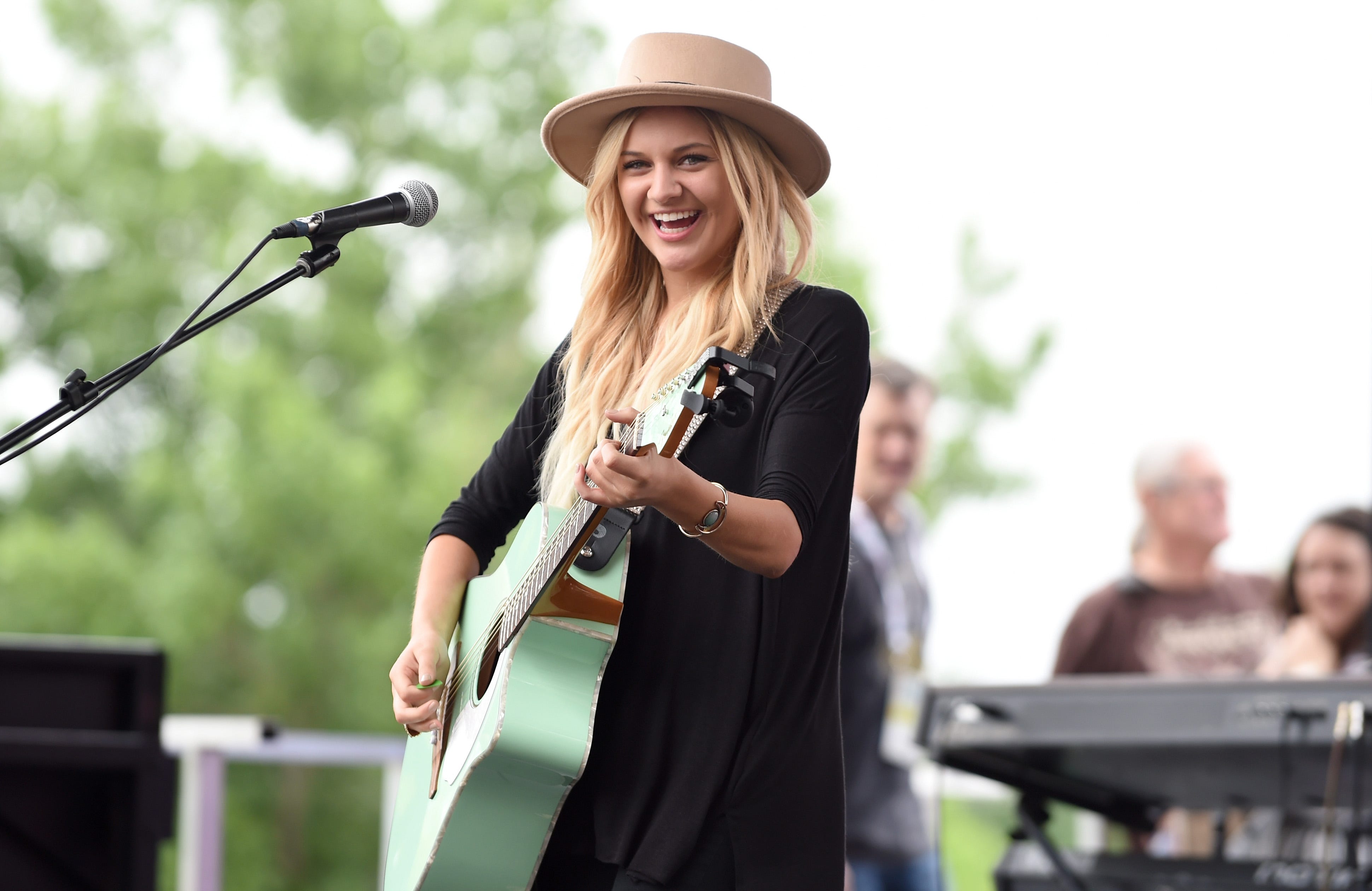 kelsea ballerini plays guitar and smiles at the camera while on a stage, she wears a tan hat and black dress, a microphone on a stand is on the left