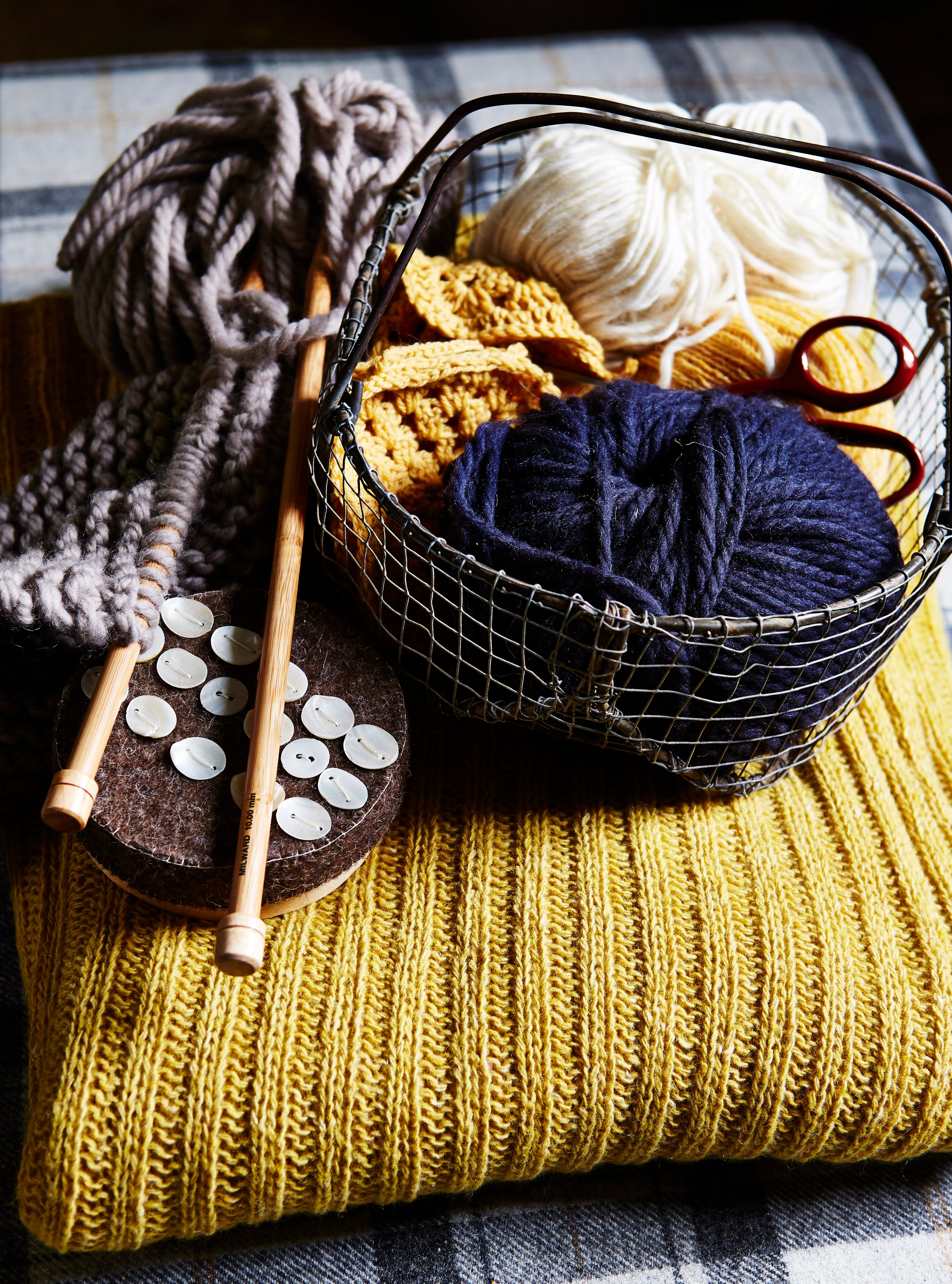 balls of blue and white wool in small wire basket, wooden knitting needles, buttons, placed on yellow ribbed knitted fabric, texture