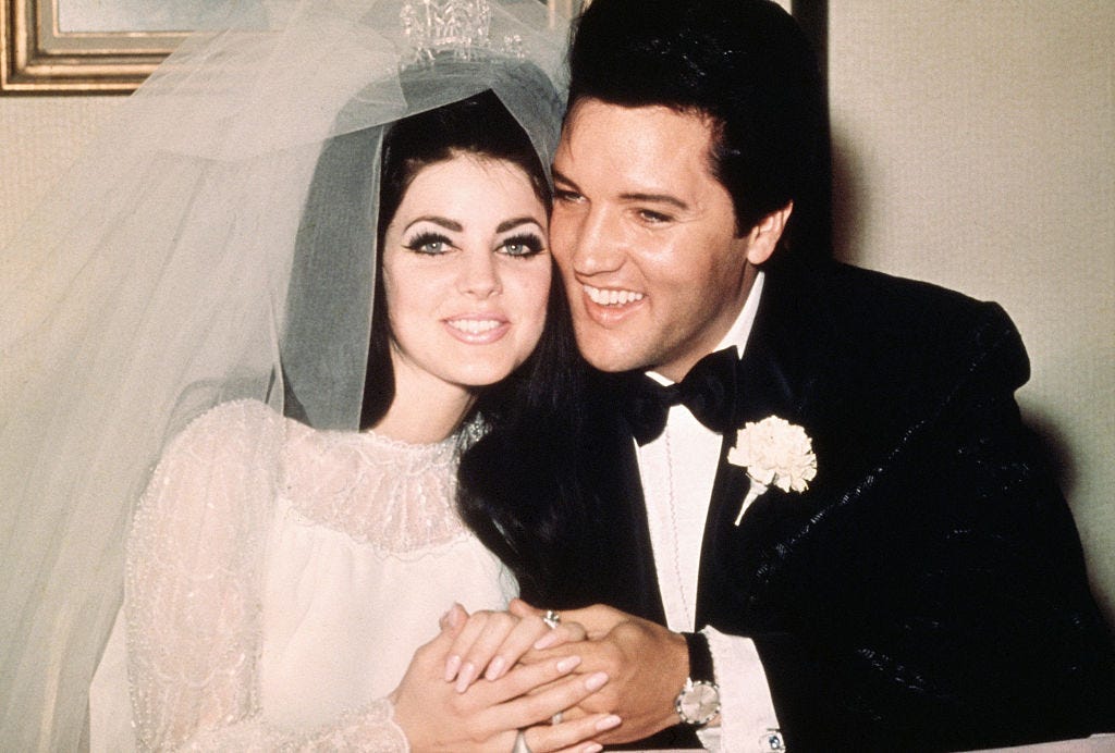 priscilla and elvis presley sit smiling and holding hands, she wears a wedding dress, veil and tiara, he wears a black suit with a bow tie and a carnation on his lapel