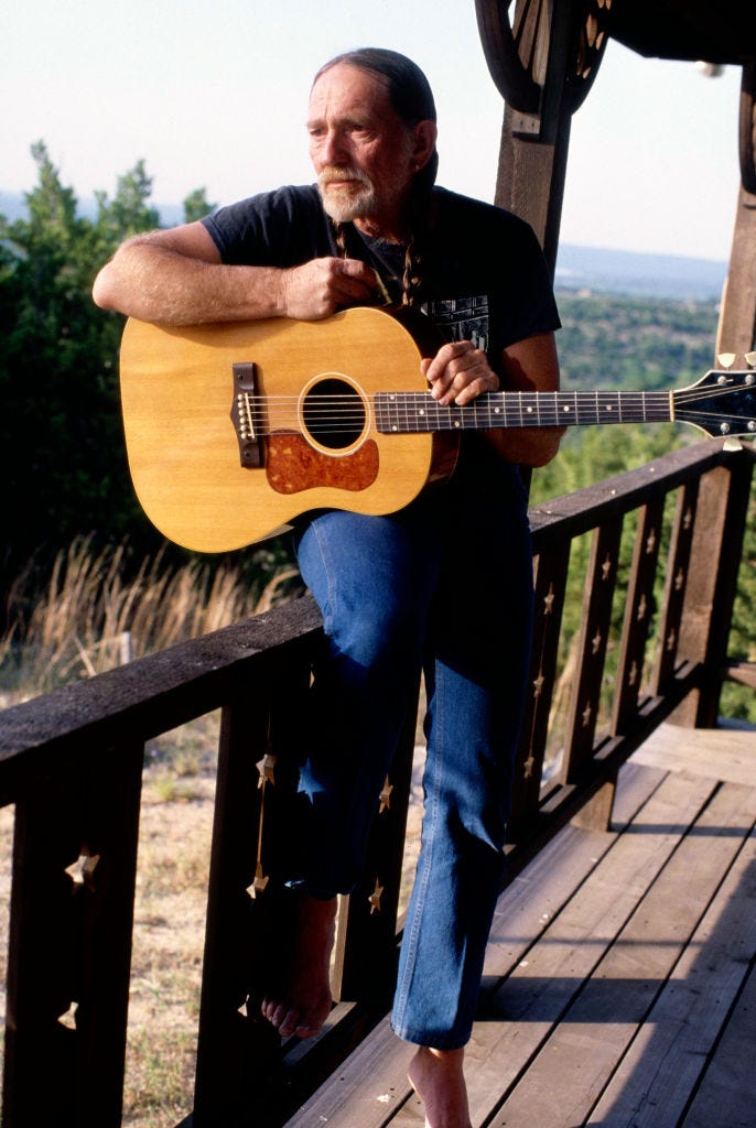 willie nelson standing against a fence and holding a guitar