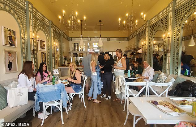 The inside of the restaurant features white tables on hardwood floors with opulent chandeliers overhead and Parisian-style artwork set across from a wall of green-framed mirrors