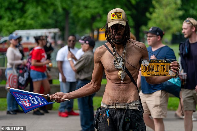 A shirtless Trump supporter with jeans, a flag and a Trump license plate walks around outside the rally