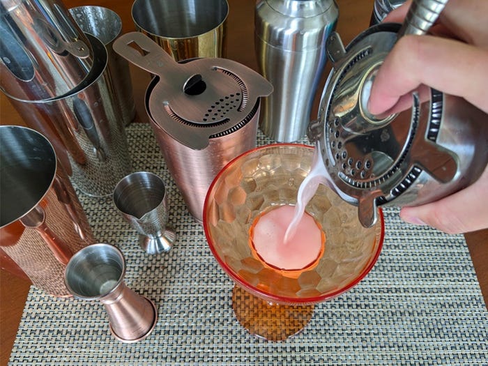 A hand pouring a drink into a cocktail glass with various cocktail shakers in the background.