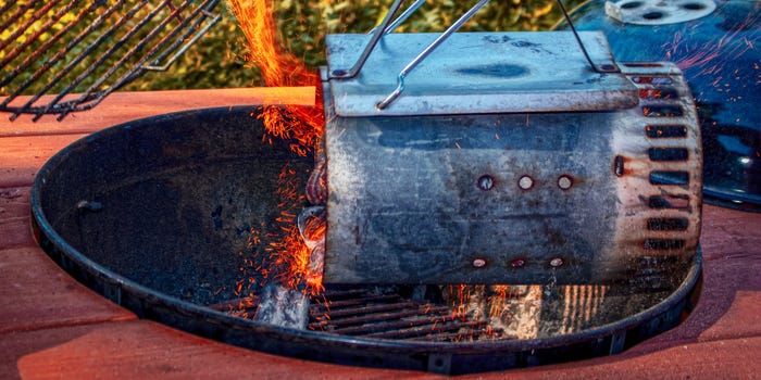 A smudged and well-used charcoal chimney starter pouring lit coals into a charcoal grill.