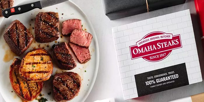 Omaha Steaks box with cooked hamburgers, cuts of beef, and chicken on a table.