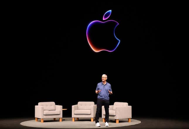 Tim Cook talking on stage in front of three empty grey chairs with an Apple logo behind him
