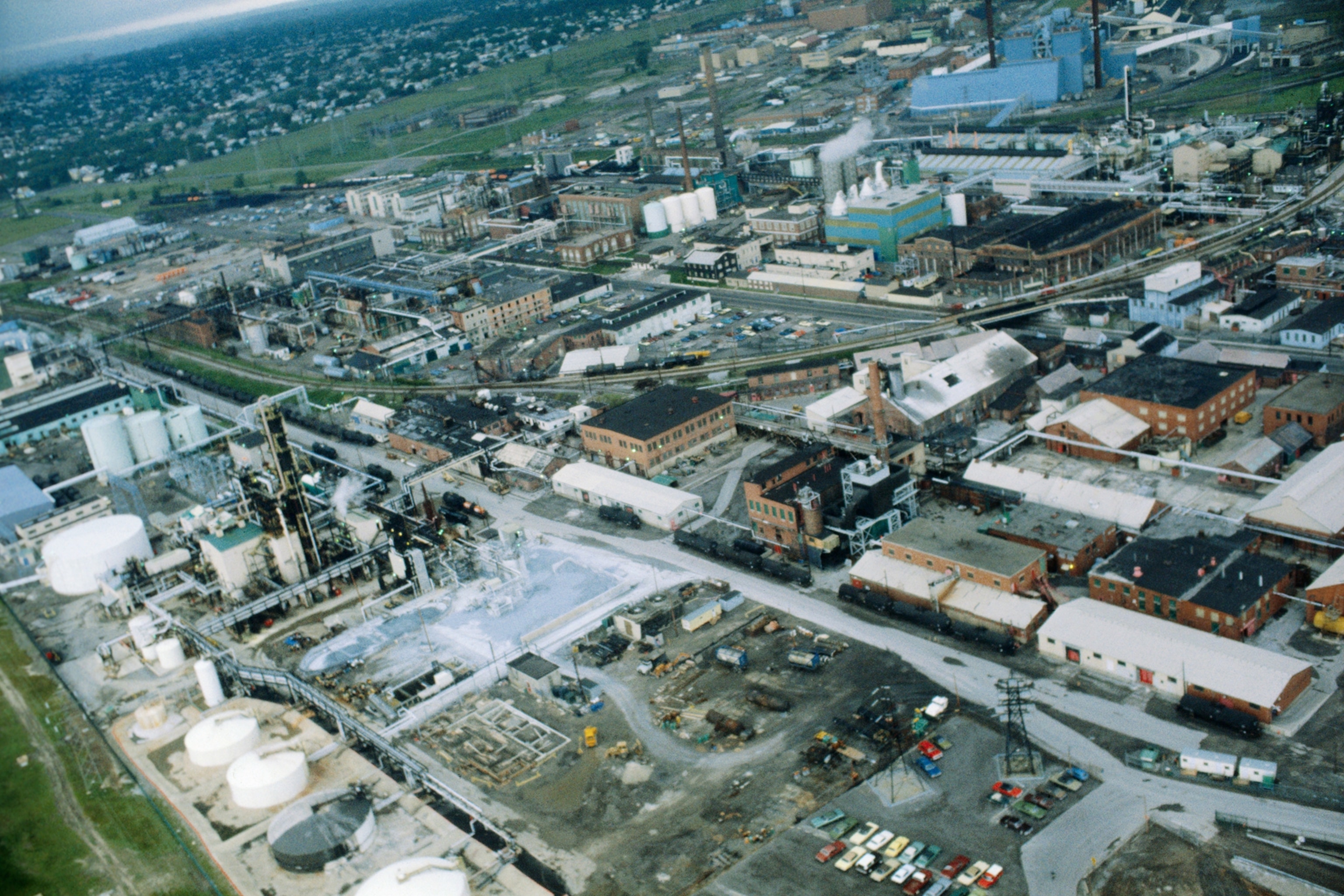An aerial view shows an industrial space, buildings, parking lots and smoke. In the background you can see the town nearby.