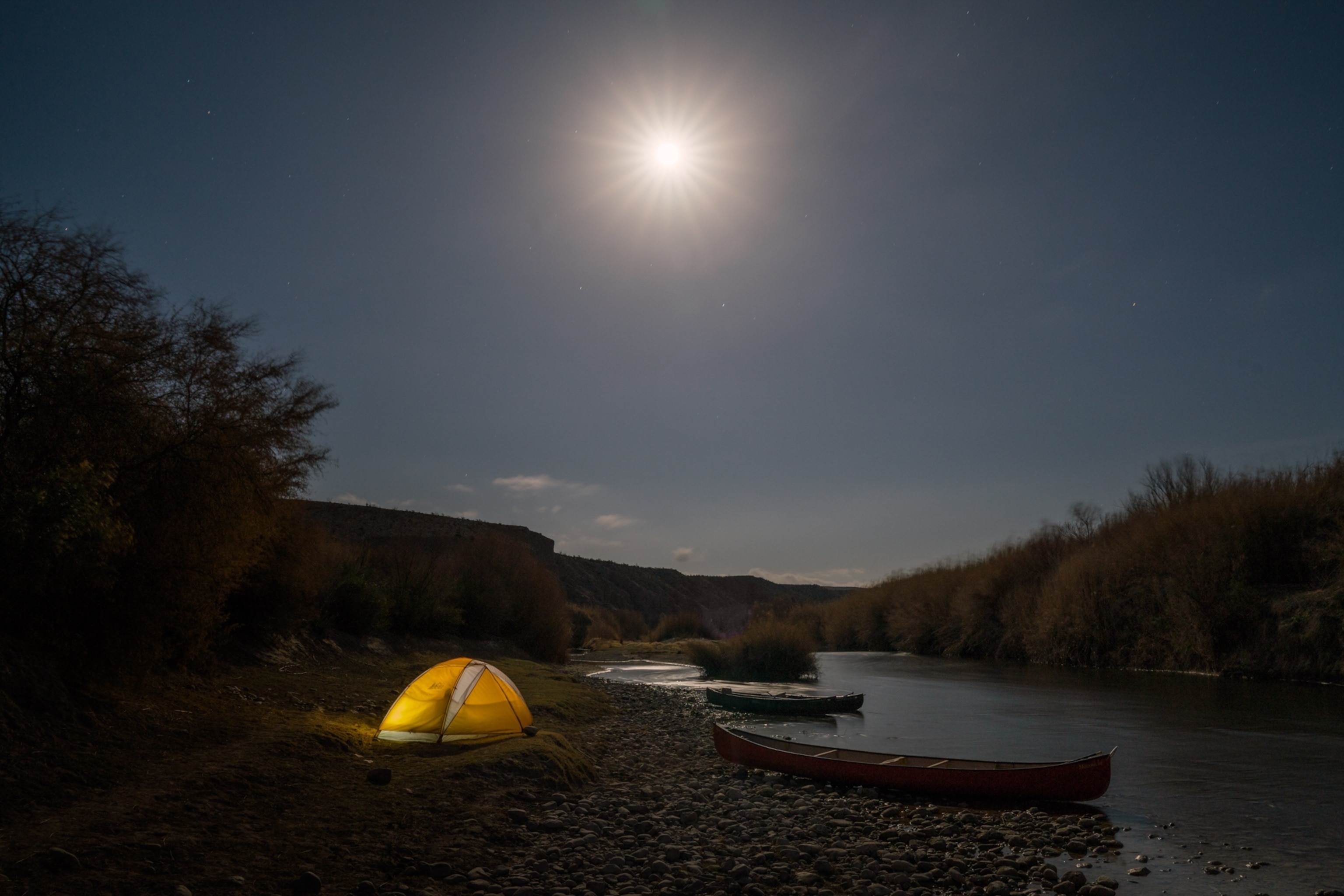 A bright moon shines above a tent and canoes camped along the river's edge of the Rio Grande.
