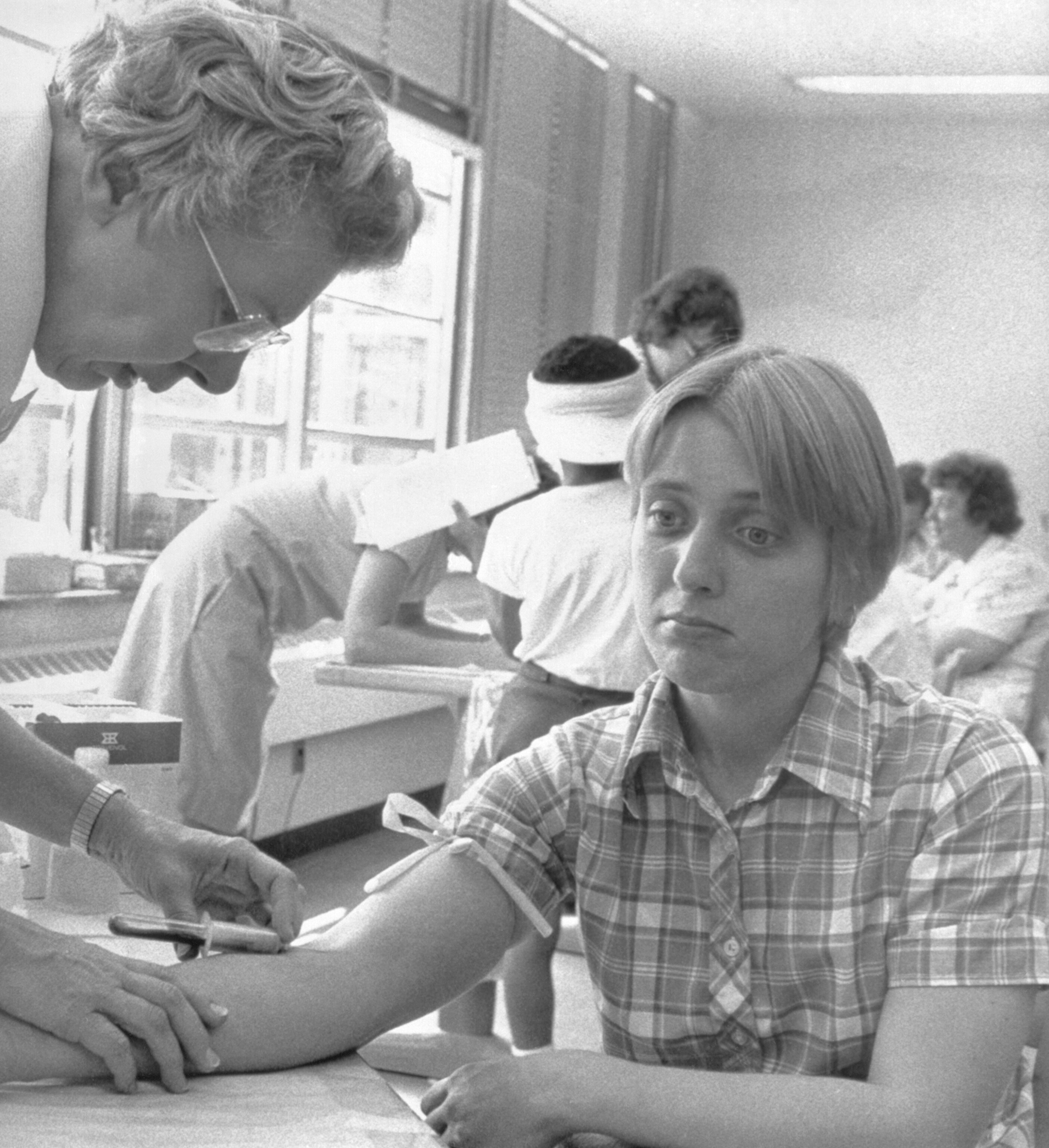 A black and white photograph of a young woman, with a solemn expression a nurse stands above her focused on pulling blood.