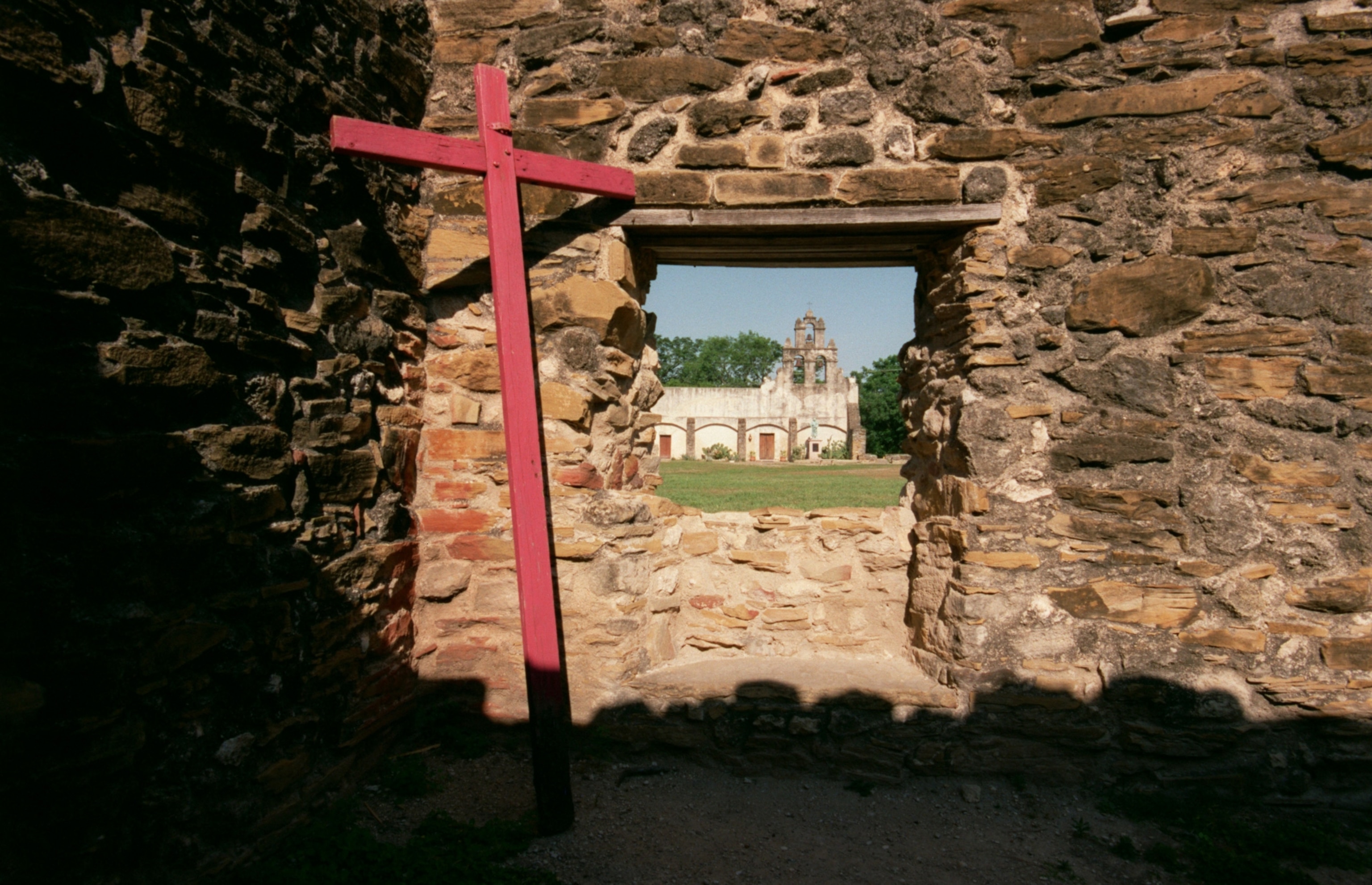 The rising church at Mission San Juan in San Antonio is seen through the window of one of the crumbling ruins across its wide plaza.