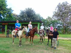 four people are riding horses in front of a green building with trees on the other side