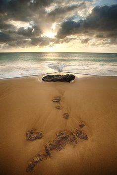 footprints in the sand on a beach near the ocean with sun shining through dark clouds