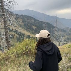 a woman standing on top of a lush green hillside next to a tall pine tree