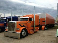 several semi trucks are lined up in a parking lot with cloudy skies behind them and one is orange