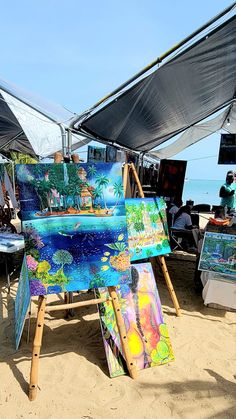 an easel is set up on the beach with paintings and umbrellas over it