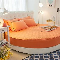 a round bed with orange sheets and pillows in a white room next to a window