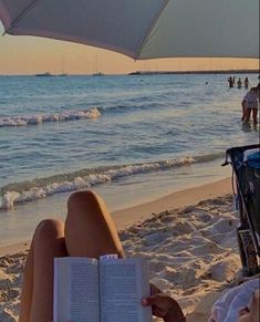 a woman reading a book on the beach while sitting under an umbrella and looking out at the ocean