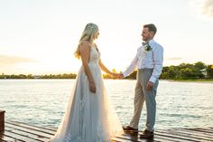 a bride and groom holding hands on a dock near the water at sunset or sunrise