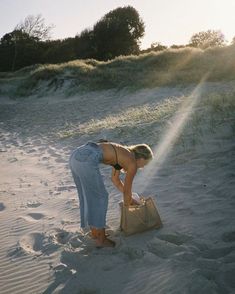 a woman bending over in the sand with a bag on her lap and sun shining behind her