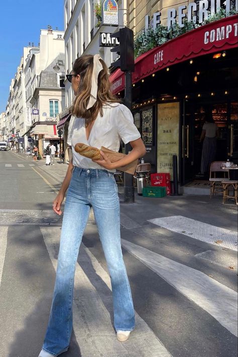 A girl is walking in the middle of the crosswalk in Paris, France. She is wearing light colored highwaisted skinny flared jeans. Her top is a wrap shirt that has a deep-V and it is fully white. Her hair is in a half-up ponytail hairstyle, and a white cream ribbon is tied to the top. Her hair is slightly wavy, and her head is looking to the side with a pair of black sunglasses. Her shoes are white and wicket brown. She is holding a baguette in her hand. The background is a corner coffee shop. Flared Jeans Summer Outfit, Bell Bottom Jeans Outfit Casual, Outfits With Light Blue Jeans, Outfits With Bell Bottom Jeans, Flare Jeans Outfit Summer, Bell Bottom Jeans Outfit Summer, Light Denim Jeans Outfit, Flared Jeans Outfit Summer, High Rise Jeans Outfit