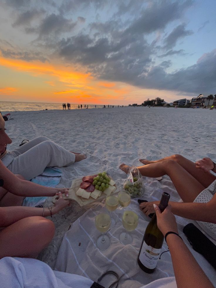 three women sitting on the beach drinking wine and having a picnic with their friends at sunset