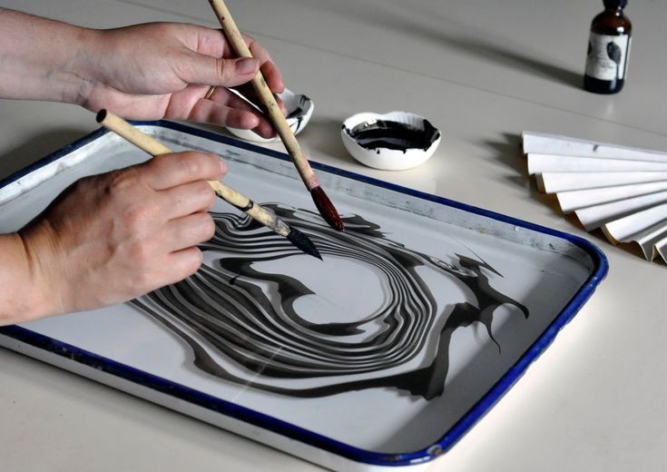 two hands holding paintbrushes over a tray with black and white designs on it