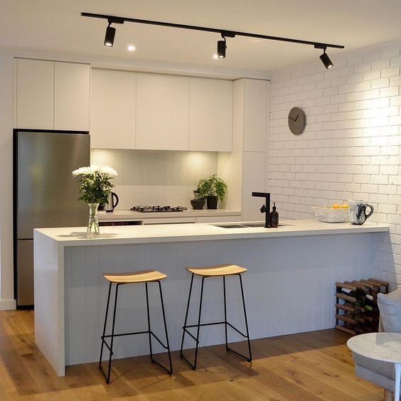two stools sit in front of an island with white countertops and cabinets, while the kitchen is lit by recessed lighting