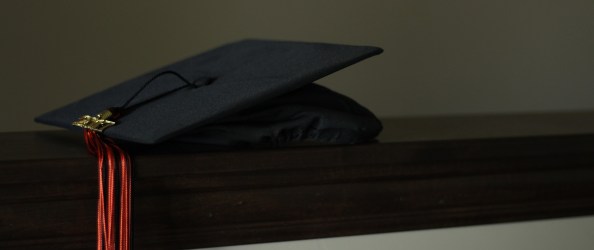 A black mortarboard with a red tassel rests on a brown banister.