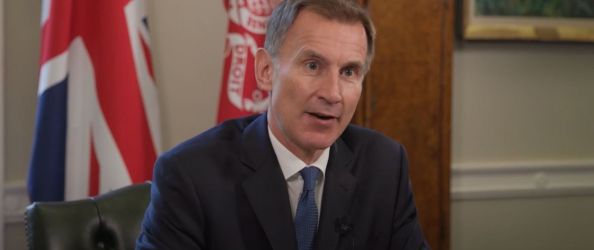 Jeremy Hunt, a white man with short brown hair and a navy suit and tie, sits in front of a desk and reads a statement. The red treasury flag and Union Jack are on poles behind him.