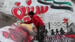 A man draws a slogan in Arabic reading "we still want freedom" as part of a graffiti mural commemorating the ninth anniversary of uprising that led to the Syrian conflict (March 15), showing a dove holding an olive branch in its beak flying over a Syrian opposition flag in the shape of the eastern Arabic numeral "9" while being targeted by the silhouette of a military aircraft with the Arabic word "years" below as silhouettes of children stand by a border fence and a tent with the letters "UN", drawn on the collapsed roof of a heavily damaged building in the town of Binnish in the northwestern Idlib province on March 13, 2020. (