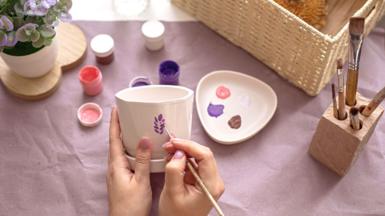 A person paints a purple flower on a cup.