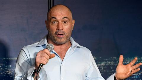 Joe Rogan performs at a comedy show, holding a mic and raising his eyebrows at the audience
