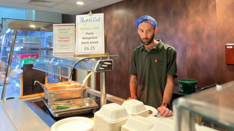 Jordan stands behind a counter where you serve food. He's wearing a green T-shirt and a blue hair net. He's next to a sign with the cafe saying it's serving pasta bolognese and garlic bread