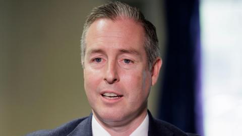 Paul Givan - a man with short hair is pictured from the shoulders up is wearing a dark suit jacket and a white collared shirt. He has his mouth open as he looks beyond the camera.