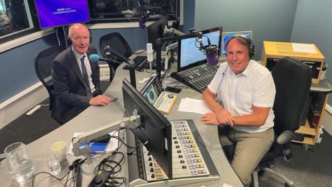 Darryl Preston is sitting at a blue microphone in a radio studio, opposite Chris Mann wearing a white shirt and beige trousers with two computer screens and two fader decks in front of him. Both are smiling at the camera