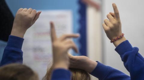 Children's hands raised in a classroom. They are wearing blue jumpers.