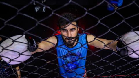 An Afghan man, looking at the camera, leans against meshed wire in a Mixed Martial Arts cage. He's wearing white boxing gloves.