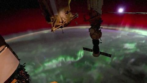 A video grab shows the northern lights (Aurora Borealis) flickering over the earth's surface with the moon setting in the background, and parts of the Internation Space Station (ISS) visible in the foreground. The Earth's curvature can be seen as it rotates