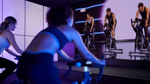 Two women on stationary exercise bikes pointing at a screen of people also on exercise bikes