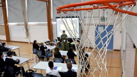 A picture of a basketball hoop in Myton School's sports hall. In the background, out of focus, a group of about a dozen students are learning at foldable desks.