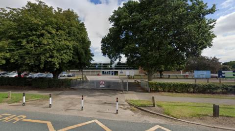 A road showing yellow zig zap lines in front of black and white waist-high barriers with red lights on. Behind is a long grey gate surrounded by green bushes leading to the school. Holderness Academy is in the distance with a car park to the left. Two big green trees are in front of the school.