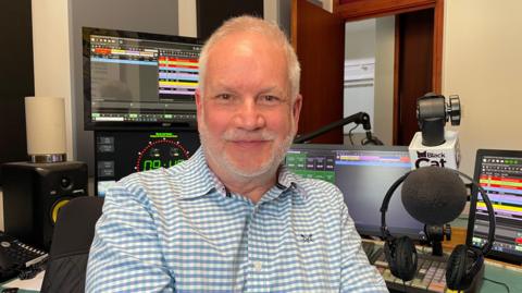 A man, Ste Greenall, sits smiling. He's in front of a radio desk, with headphones to the side of him