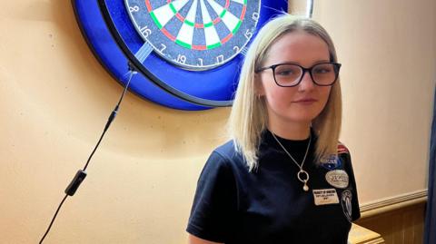 Girl wearing blue darts top poses with arms folded in front of darts board