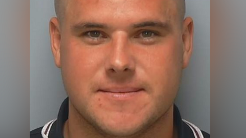 A man with hair shaven at the sides smiles at the camera in a police photo