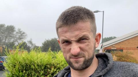 Jake Crowder, who has short hair and a beard, looks into the camera. He is wearing a grey hooded top and standing in front of a hedge