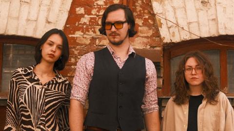 Two women stand on either side of a tall man in front of a redbrick building. The man wears a button-up vest over a floral patterned shirt, sleeves rolled up. His combover hairstyle and sunset-tinted glasses complete his 1970s look. The woman on his left has a short black bob haircut and wears a blouse with black and white zebra print. The woman on the man's right has shoulder-length, curly hair and wears glasses. She's wearing a cream-coloured, oversized shirt over a black outfit.