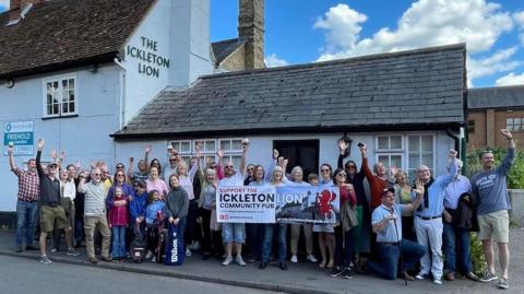 About 30 people on the pavement outside The Ickleton Lion pub holding a banner with their details on to save the pub.